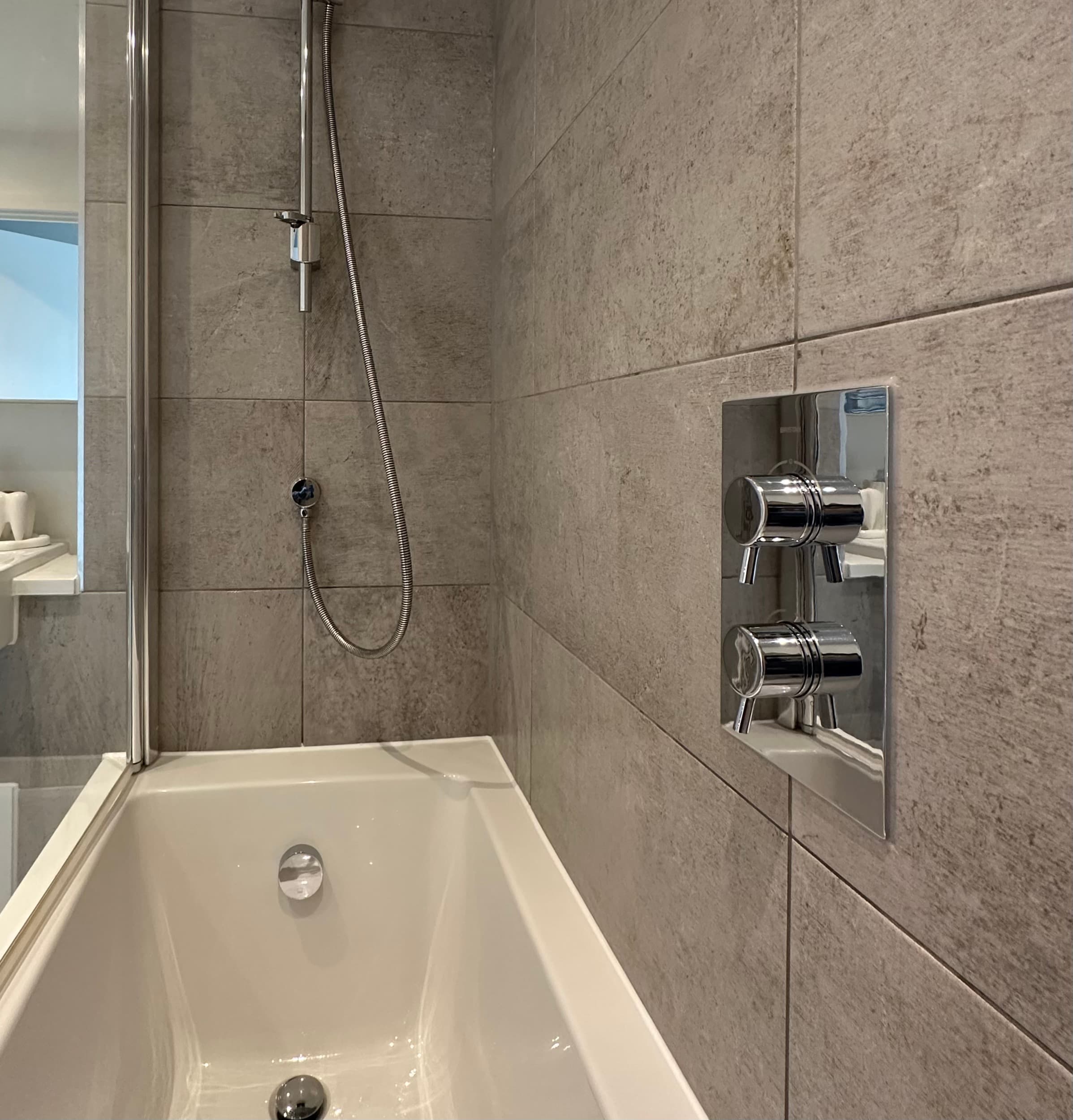 White bathtub against grey stone tiles with chrome shower hose and wall-mounted mixer controls.